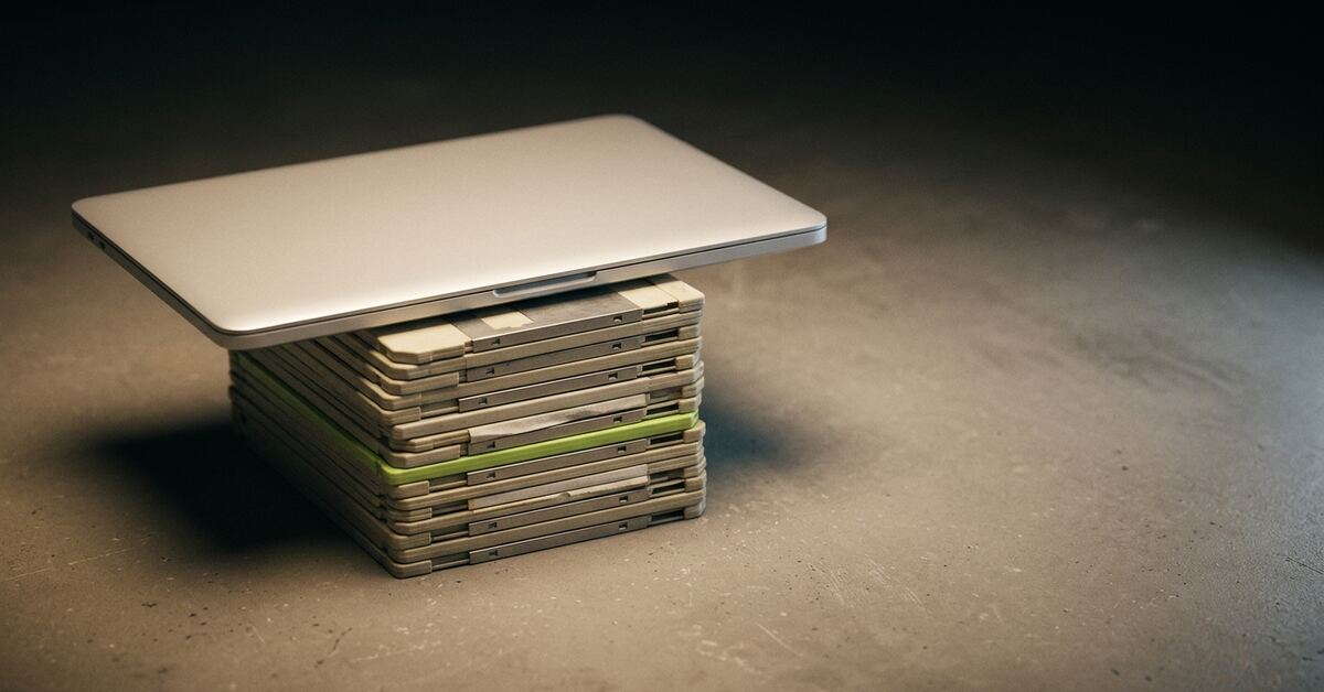 A thin modern laptop resting flat on top of a neat stack of aged beige floppy disks, one of them lime green, on a workbench.