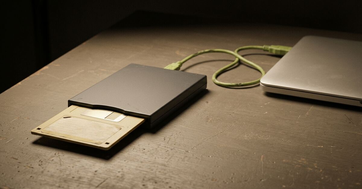 An aged beige floppy disk half-inserted into a slim USB floppy drive, its lime-green cable curving across a workbench towards the corner of a closed modern laptop.