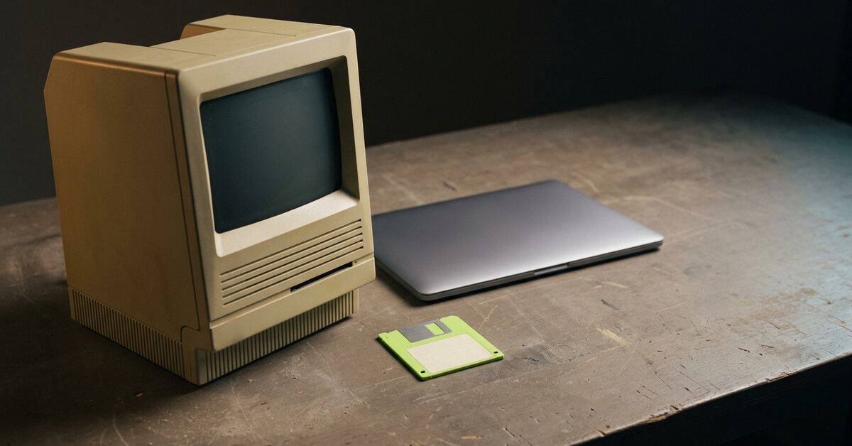 A beige 1980s compact computer with its screen dark, beside a closed modern laptop on a workbench, with a lime-green floppy disk lying between them.